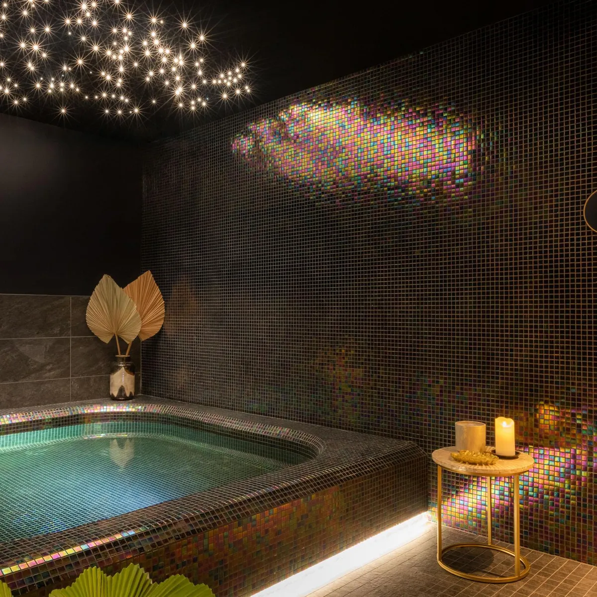 Bathtub built into a wall covered with iridescent mosaic tiles, lit by stars on the ceiling and a candle on a low table.