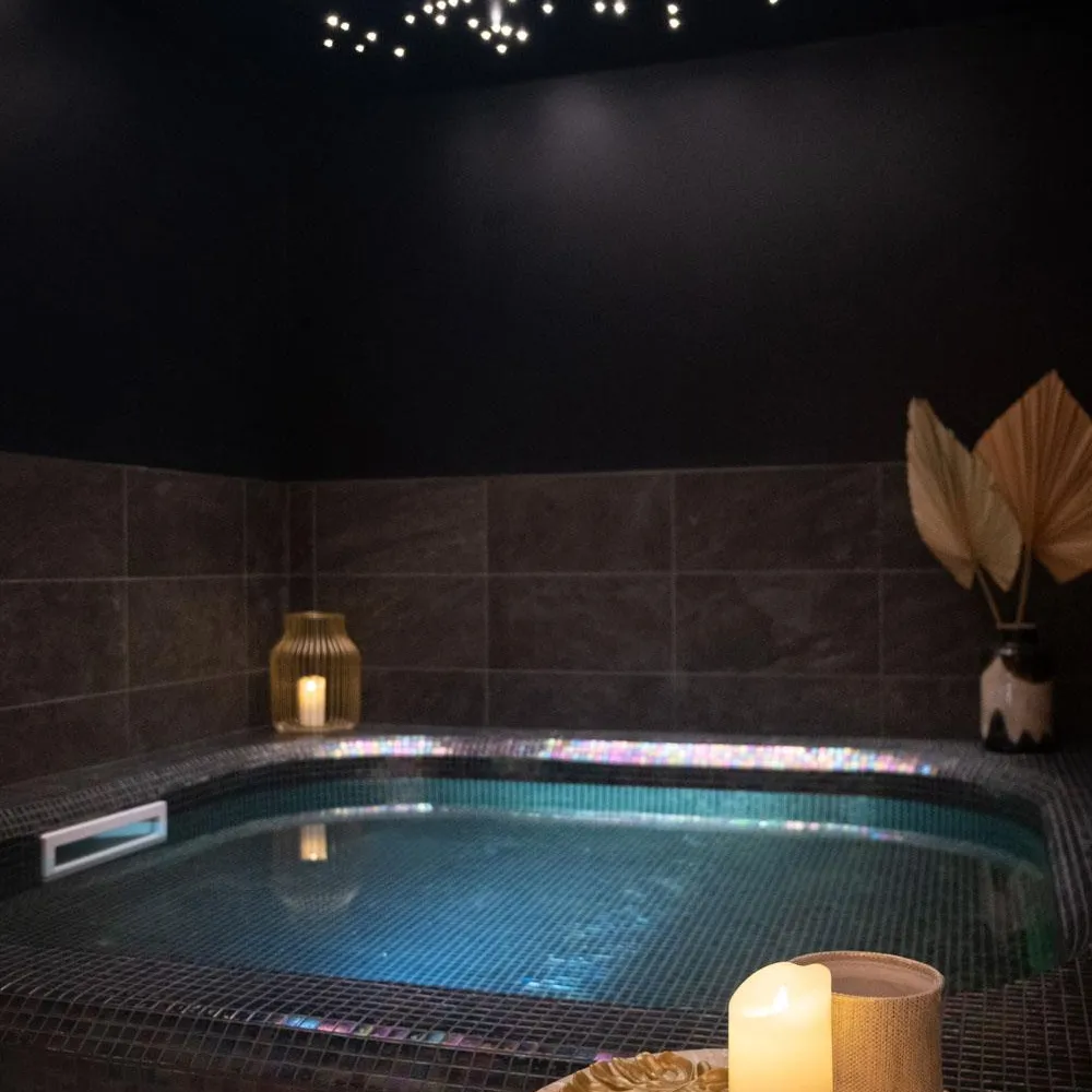 Indoor pool illuminated with underwater lights, surrounded by dark tiles, and a table with a lit candle and a vase containing a decorative leaf