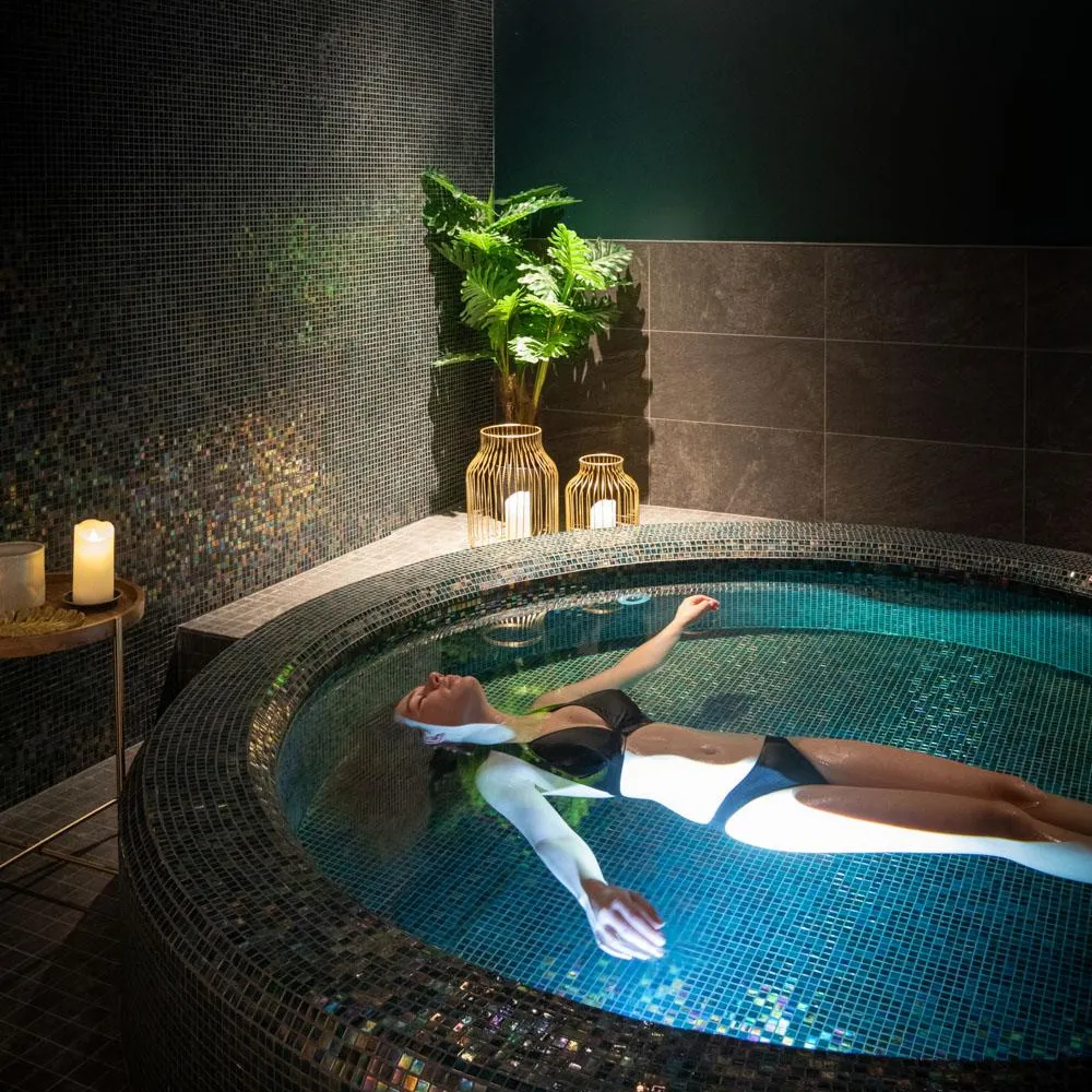 A person in a swimsuit floats in a hot tub surrounded by mosaics, with candles and plants in the background.