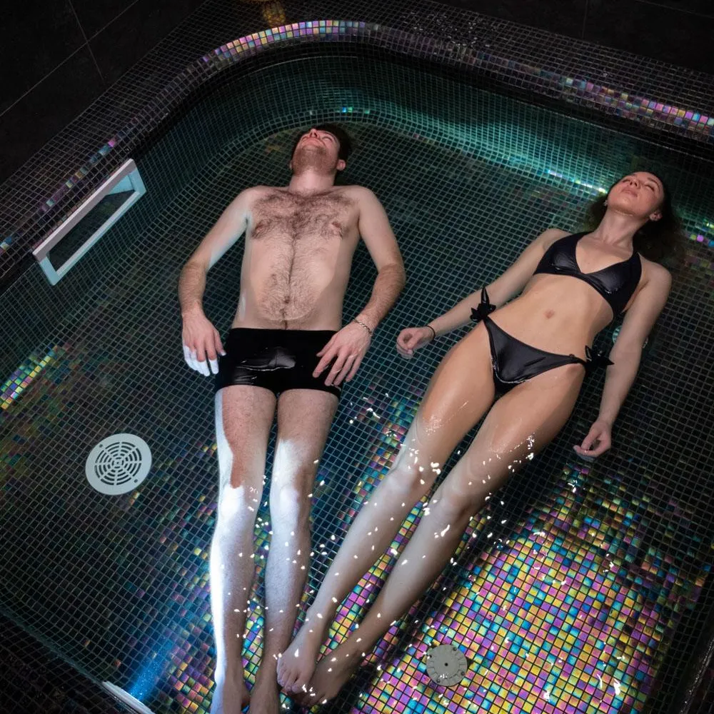 Two people lying in a hot tub with water jets, the man shirtless and the woman in a black swimsuit.