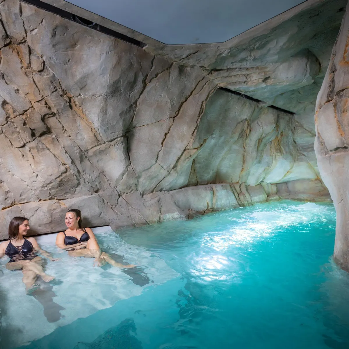 Zwei Personen sitzen in einem türkisfarbenen Wasserbecken, das von Felswänden umgeben ist.