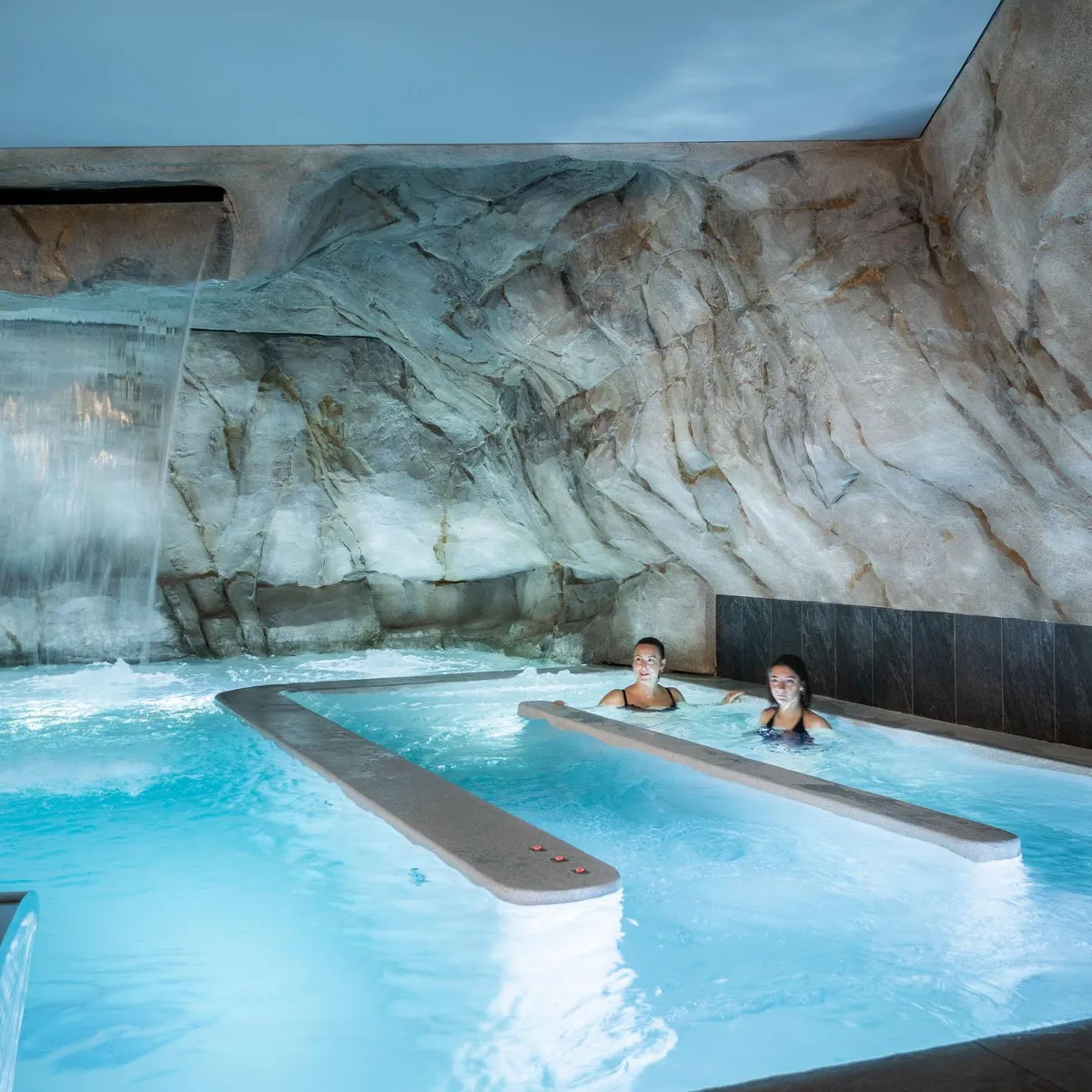 Zwei Personen schwimmen in einem Hallenbad mit einem Wasserfall und künstlichen Felsen im Hintergrund.