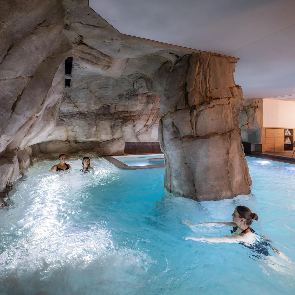 Drei Personen schwimmen in einem Innenpool mit felsigen Wänden, wobei ein Ruheraum im Hintergrund sichtbar ist.