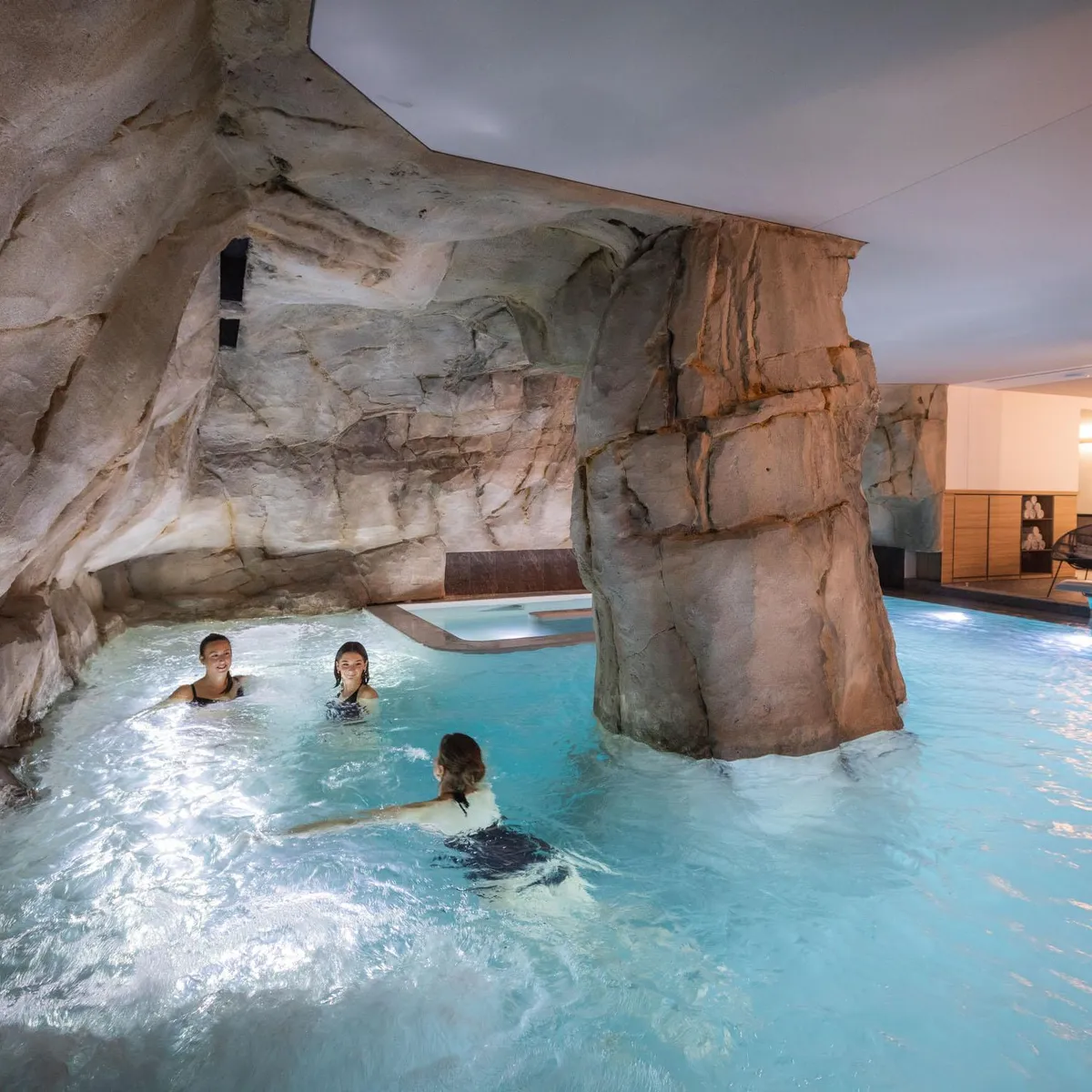Drei Personen schwimmen in einem Innenpool, der von künstlichen Felswänden umgeben ist, mit Liegestühlen und Lichtern im Hintergrund.