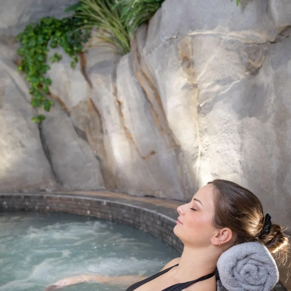 Frau in schwarzem Badeanzug liegt in einem Whirlpool mit einem grauen Handtuch hinter dem Kopf, umgeben von Felsen und Vegetation.