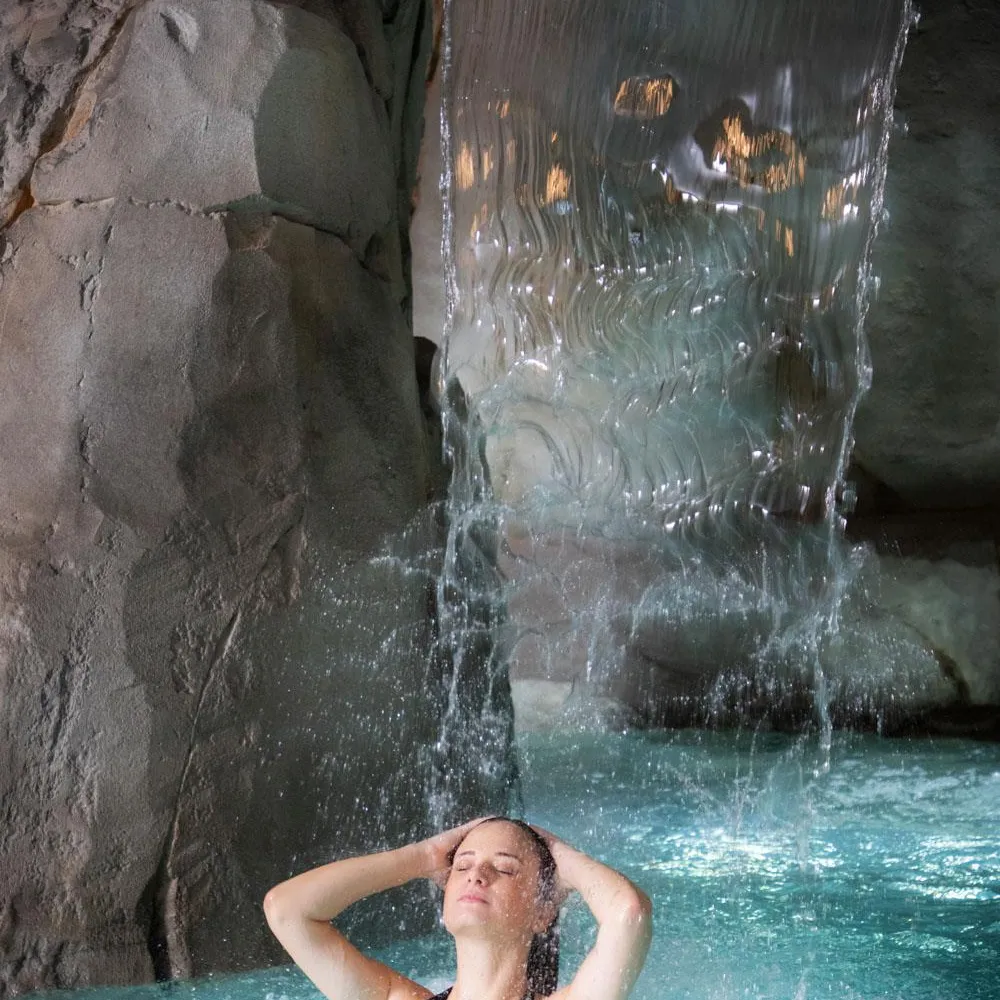 Eine Person ist in einem Becken unter einem Wasserfall eingetaucht, die Hände hinter dem Kopf, in einer felsigen Umgebung.
