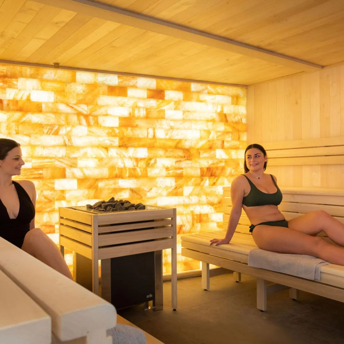 Zwei Frauen sitzen auf Holzbänken in einem Sauna, der durch eine beleuchtete Wand mit goldenen Mustern erhellt wird.
