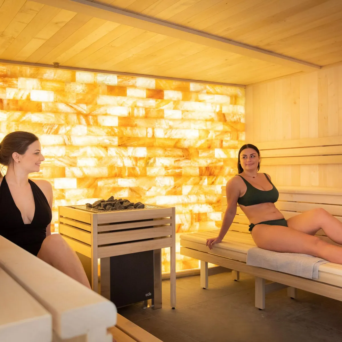 Zwei Frauen sitzen in einer Holssauna mit heißen Steinen in der Mitte und einer beleuchteten Wand im Hintergrund.