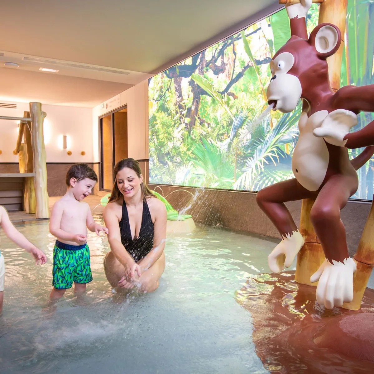 Eine Frau und zwei Kinder spielen in einem Hallenbad mit Dschungeldeko und einer Tierskulptur.