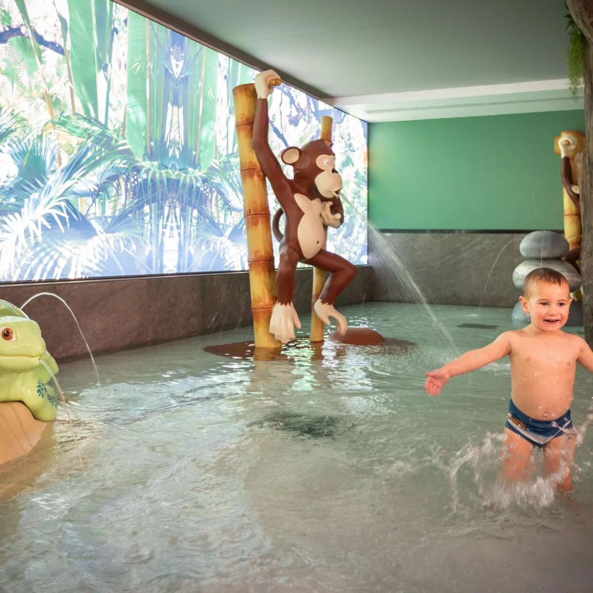 Kind spielt in einem Kinderbecken mit Tierskulpturen und Wasserfontänen, dekoriert mit Dschungelmustern