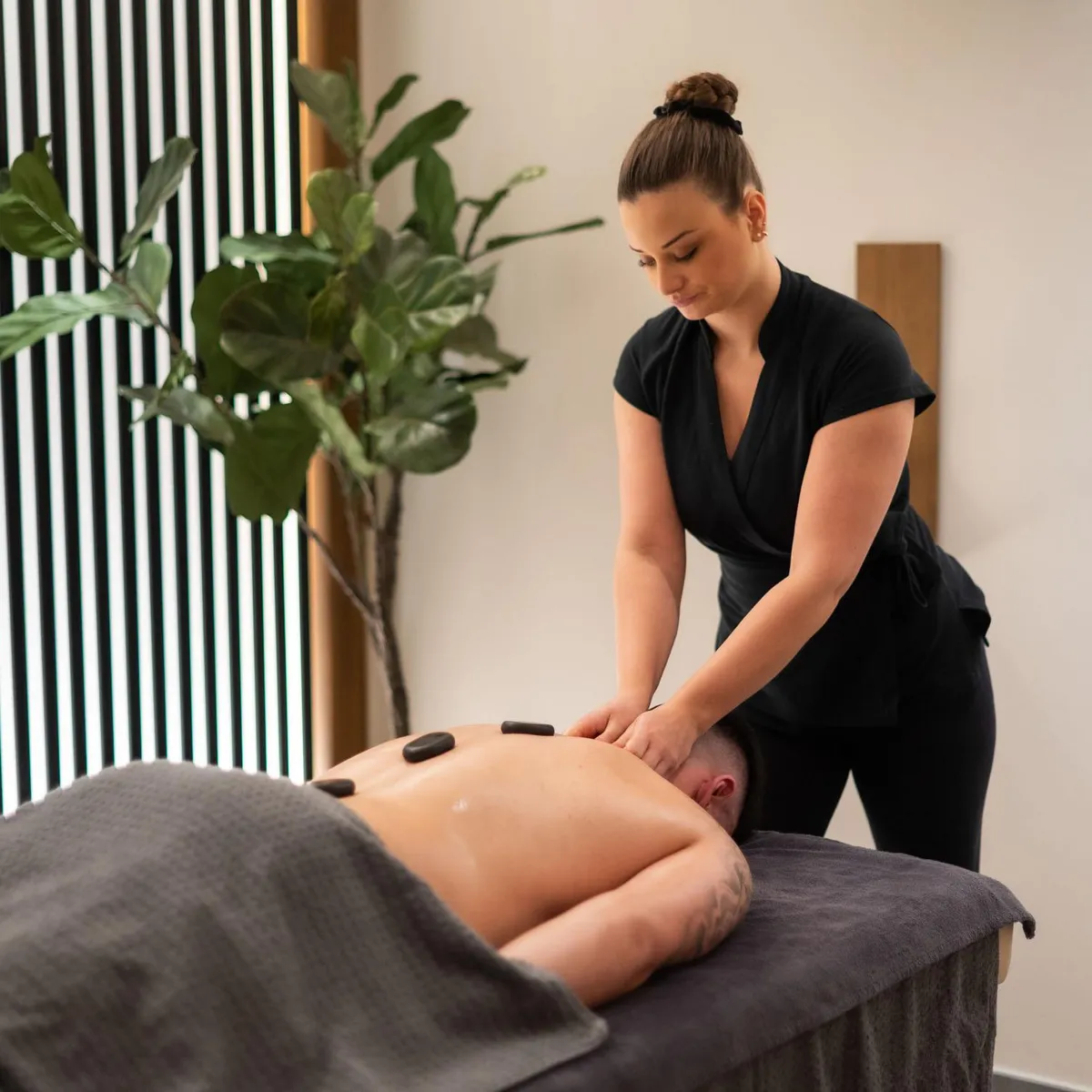 A woman in black attire is performing a massage on the back of a person lying on a table covered with a towel, with a green plant nearby.