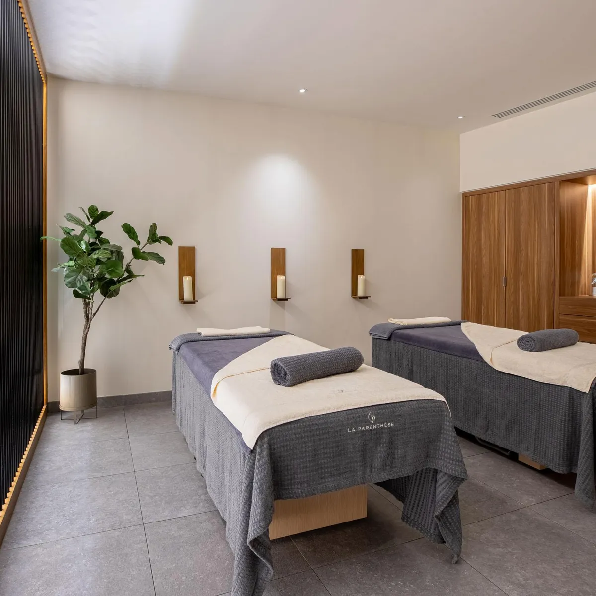 Two massage tables with gray and white towels, a green plant, and wall sconces in a room with beige walls and a gray floor.