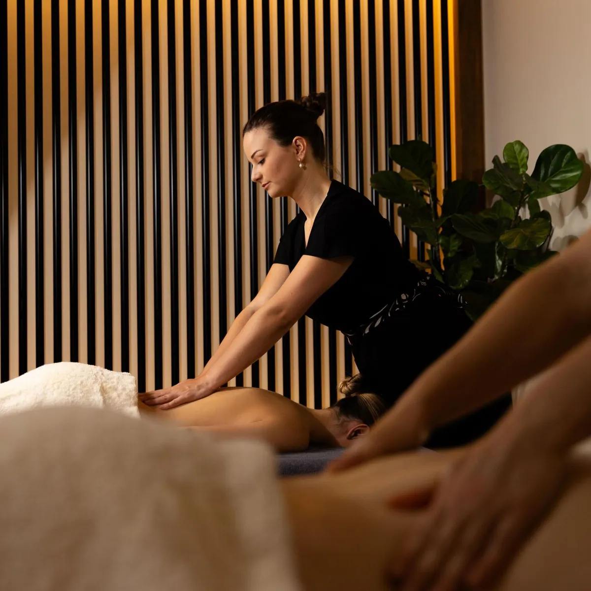 A person in black attire is giving a massage to a person lying face down on a table covered with a towel.
