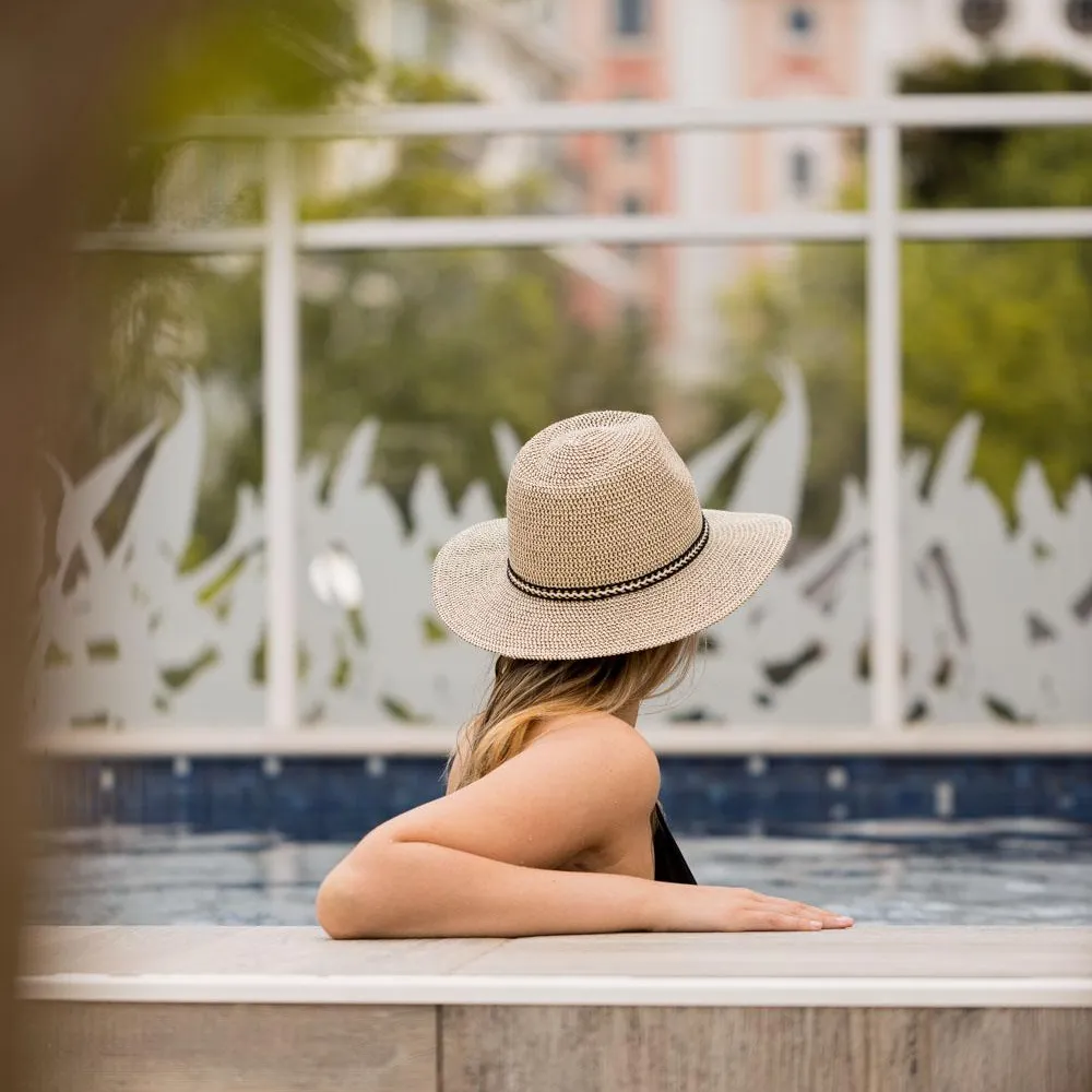 A person wearing a straw hat is sitting at the edge of a pool, with their back turned to the viewer, and a building in the background.