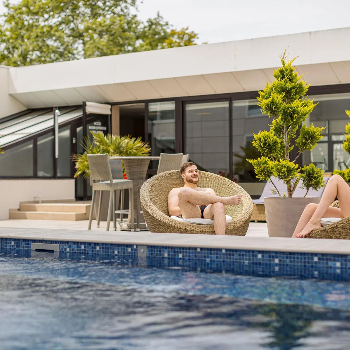 Two people sitting in wicker chairs by the edge of a pool in front of a modern building with potted plants.
