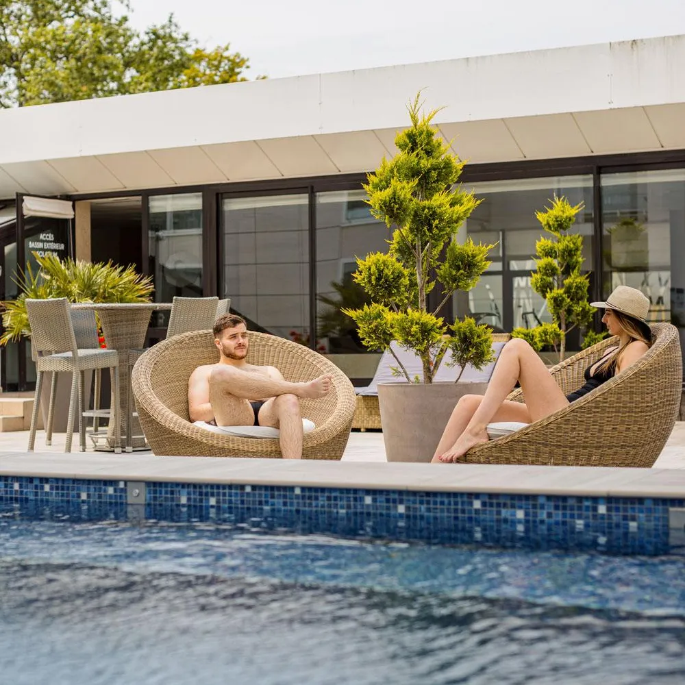 Two people sitting in wicker chairs by the edge of a pool in front of a building with large glass windows.