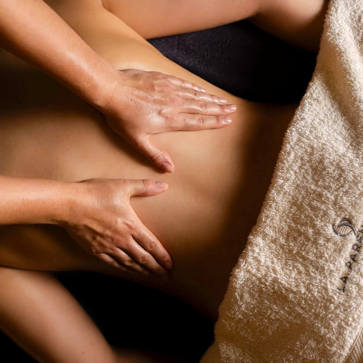 Two hands massage the abdomen of a person lying down, covered with a towel.