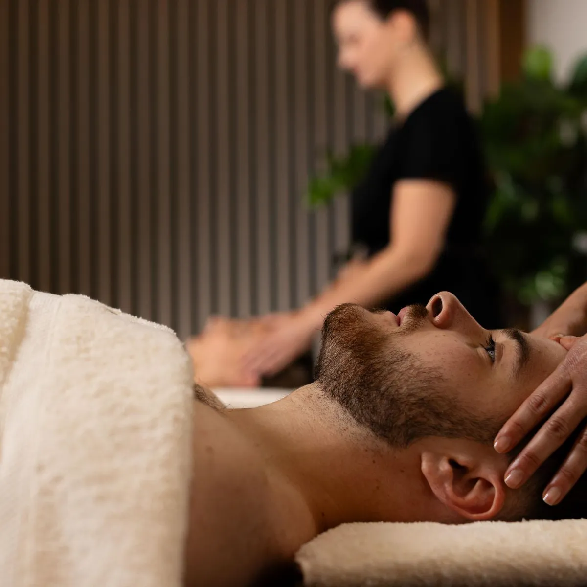 A man lying on his stomach receives a neck massage from a professional, in an indoor setting with plants in the background.