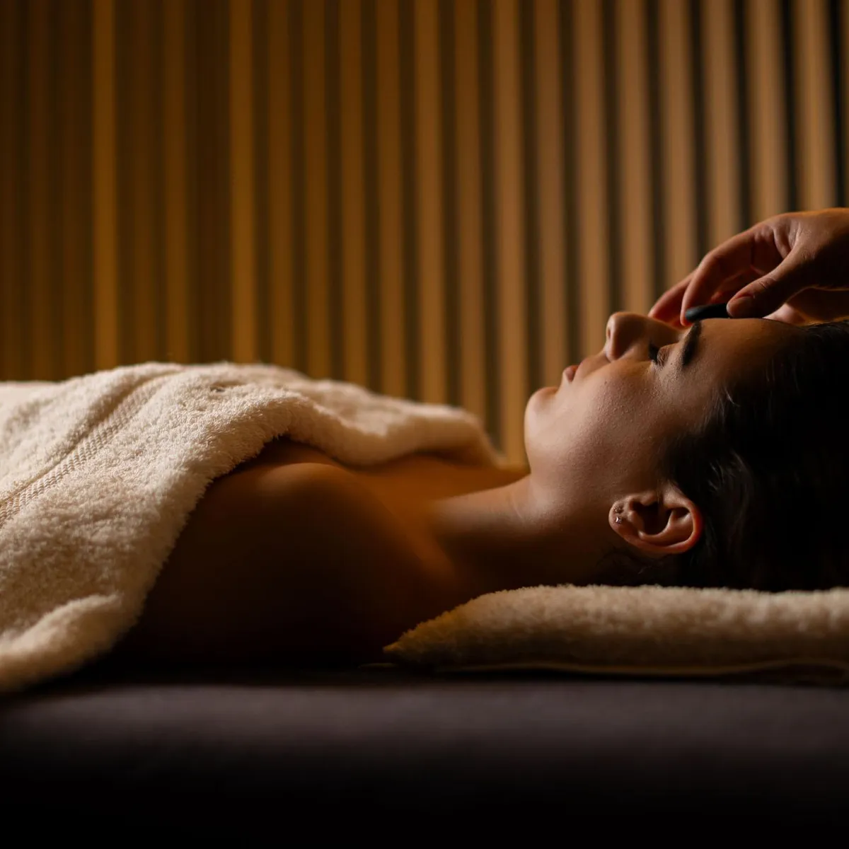 A person lying on a towel receives a facial massage in a room with striped walls.
