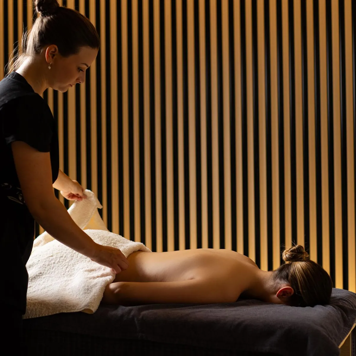 A person in black attire is giving a massage to another person lying on a table covered with towels.