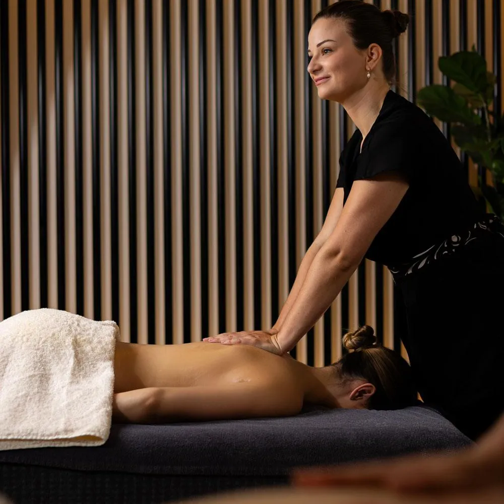 A woman in black attire is giving a massage to a person lying on a table, in a room with vertically striped walls.