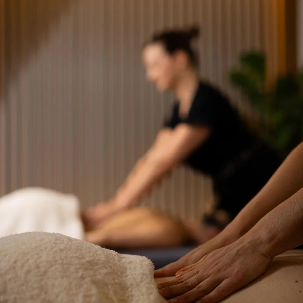 A person receives a massage on a table covered with a towel, with another person in the blurred background.