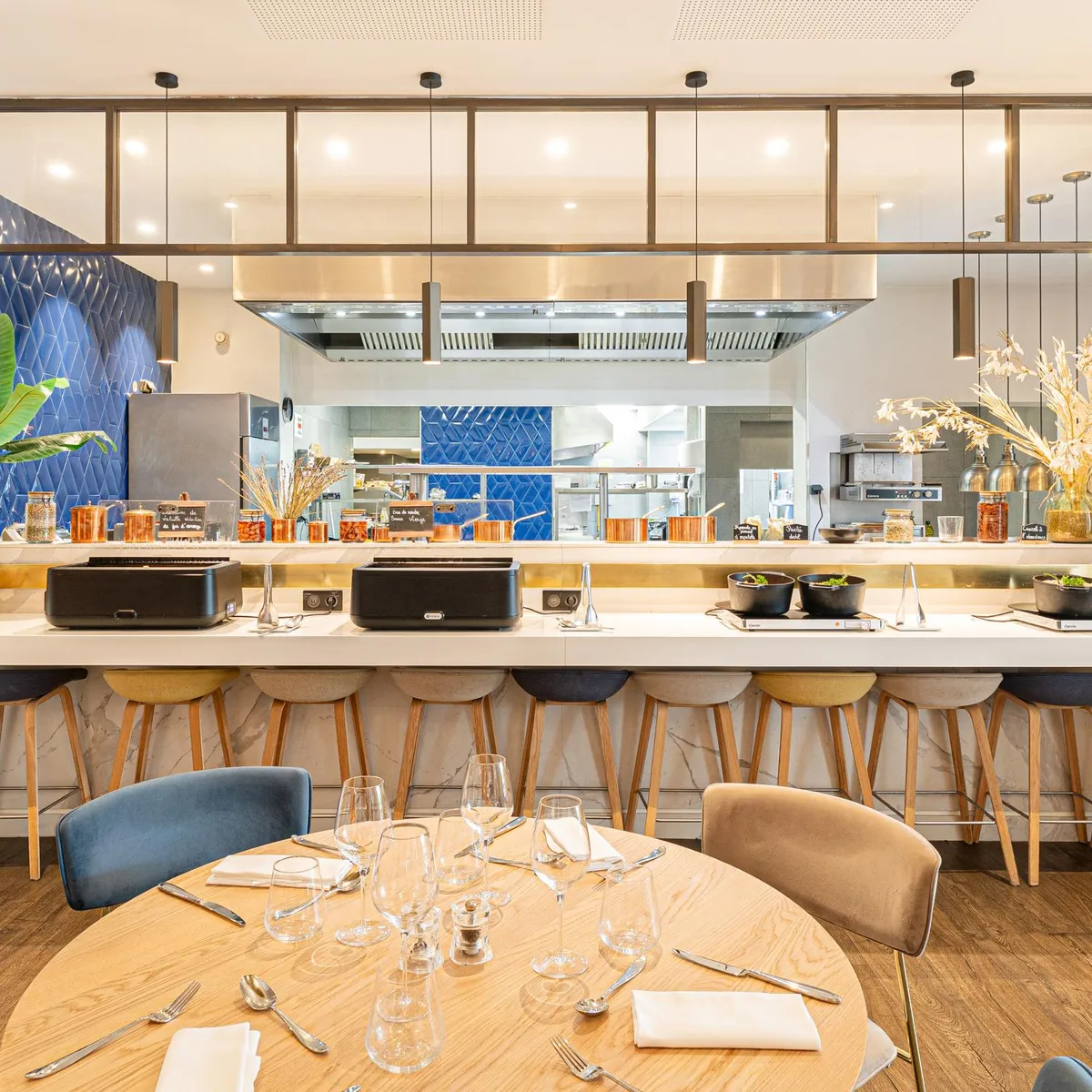 A restaurant with an open kitchen, bar stools, and a table set with glasses and cutlery.
