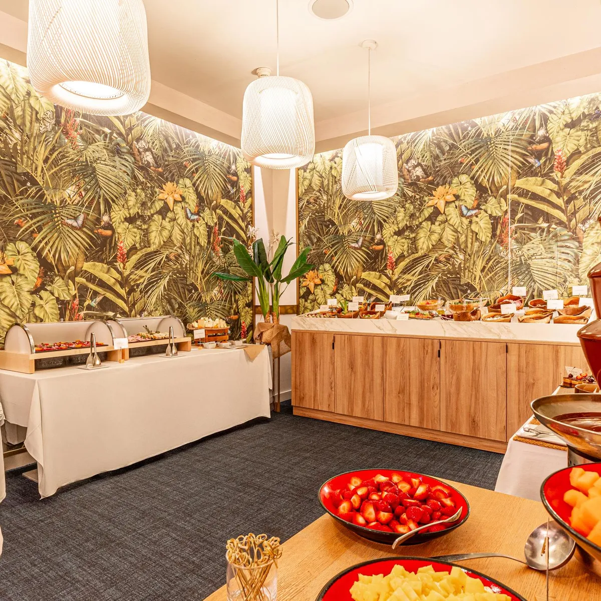 Buffet with various dishes, including a chocolate fountain, in a room decorated with tropical motifs and lit by hanging lights