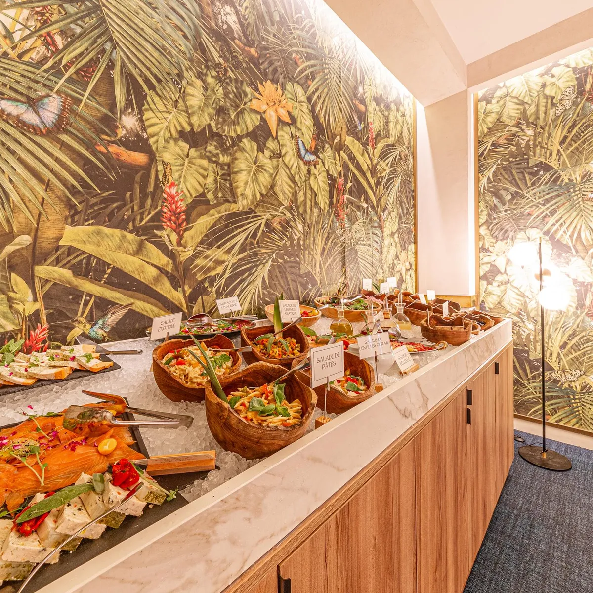 A buffet of varied dishes, including smoked salmon and salads, is presented on a table covered in ice, with walls decorated with a tropical leaf pattern.