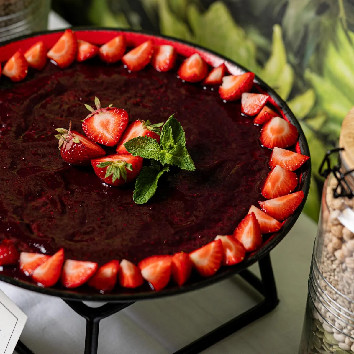 Chocolate mousse topped with strawberries and mint leaves, served on a tray with a label and surrounded by jars of legumes.
