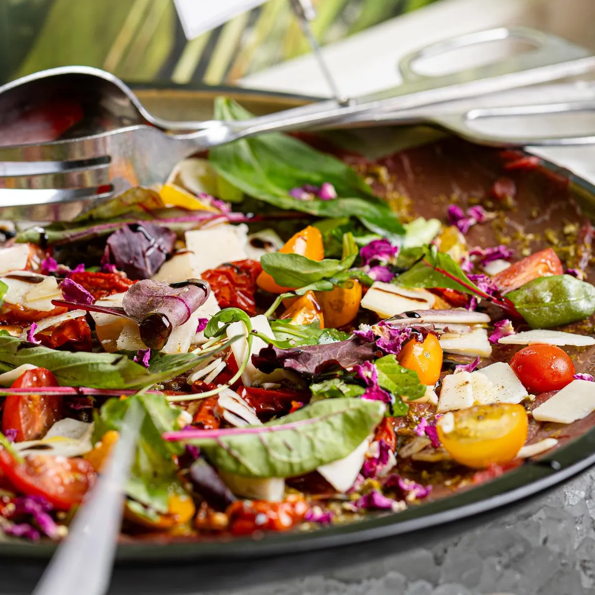 A large bowl filled with a colorful salad containing tomatoes, green vegetables, radishes, cheese cubes, and seeds, with utensils for serving.