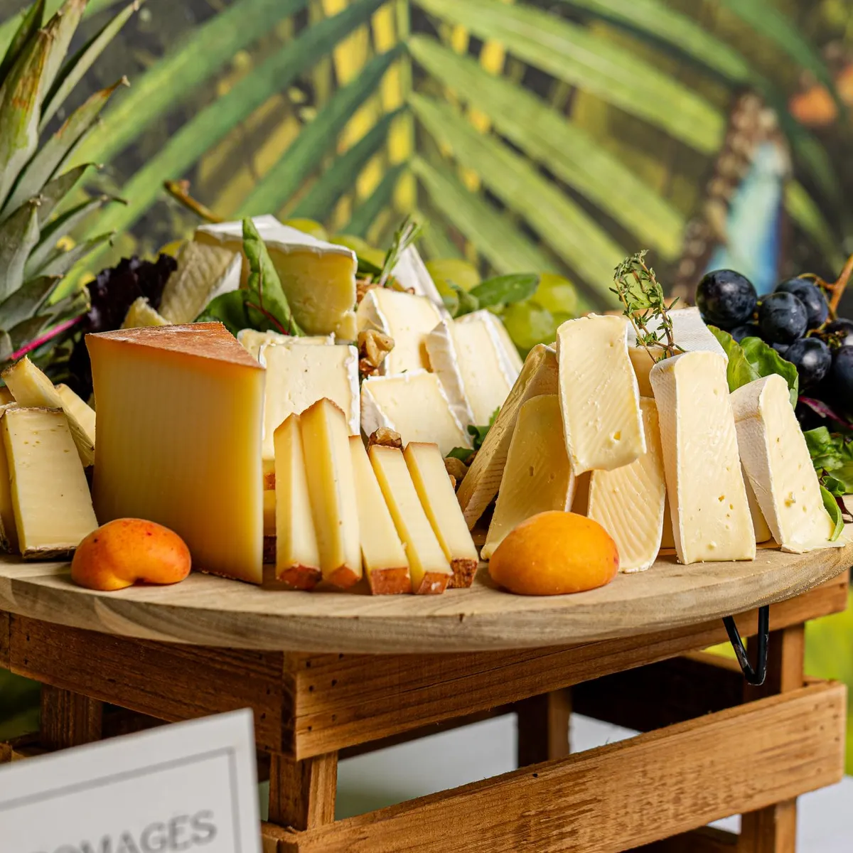 A wooden platter features various types of cheese, fruits, and basil leaves, surrounded by green plants.