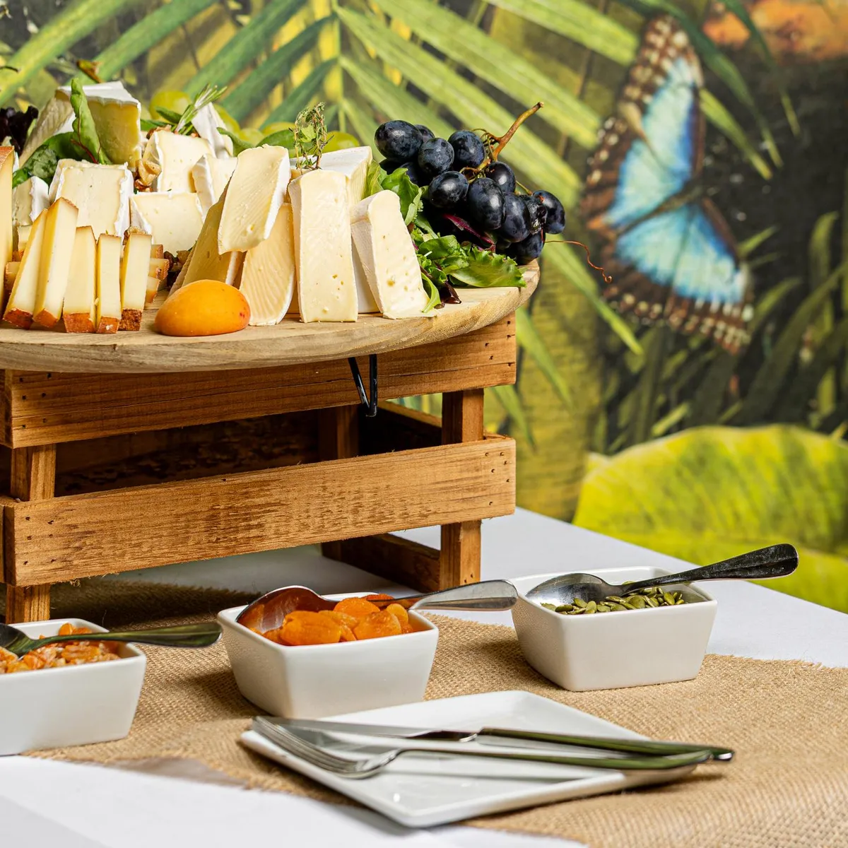A cheese and fruit platter with bowls of toppings is presented on a table covered with a jute tablecloth, with tropical decor in the background.