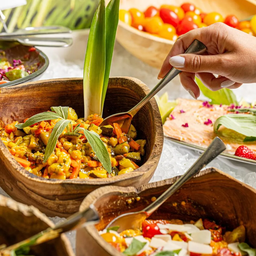 A person serves a vegetable-based dish in a wooden bowl, surrounded by similar dishes.