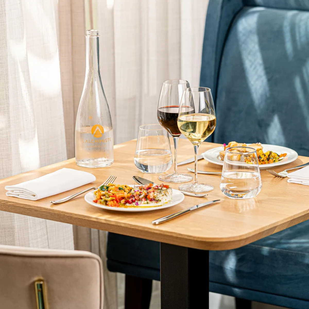 Table set with two plates of food, two wine glasses, a water bottle, and water glasses, with napkins and cutlery.