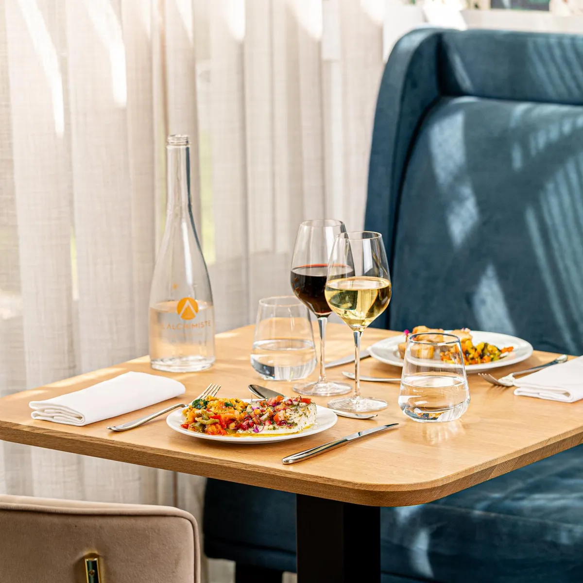 Table set with two plates of food, two wine glasses, two water glasses, a water bottle, and cutlery, in front of a blue bench and a beige chair.
