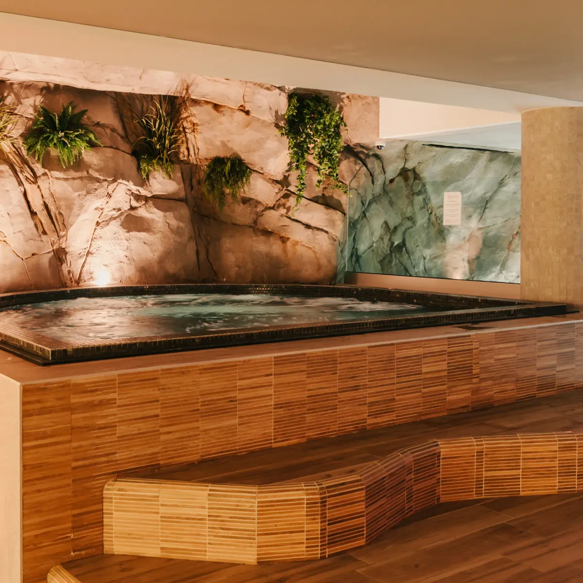 Piscine intérieure avec un mur décoré de roches et de plantes vertes, entourée de carrelages et de bancs en bois