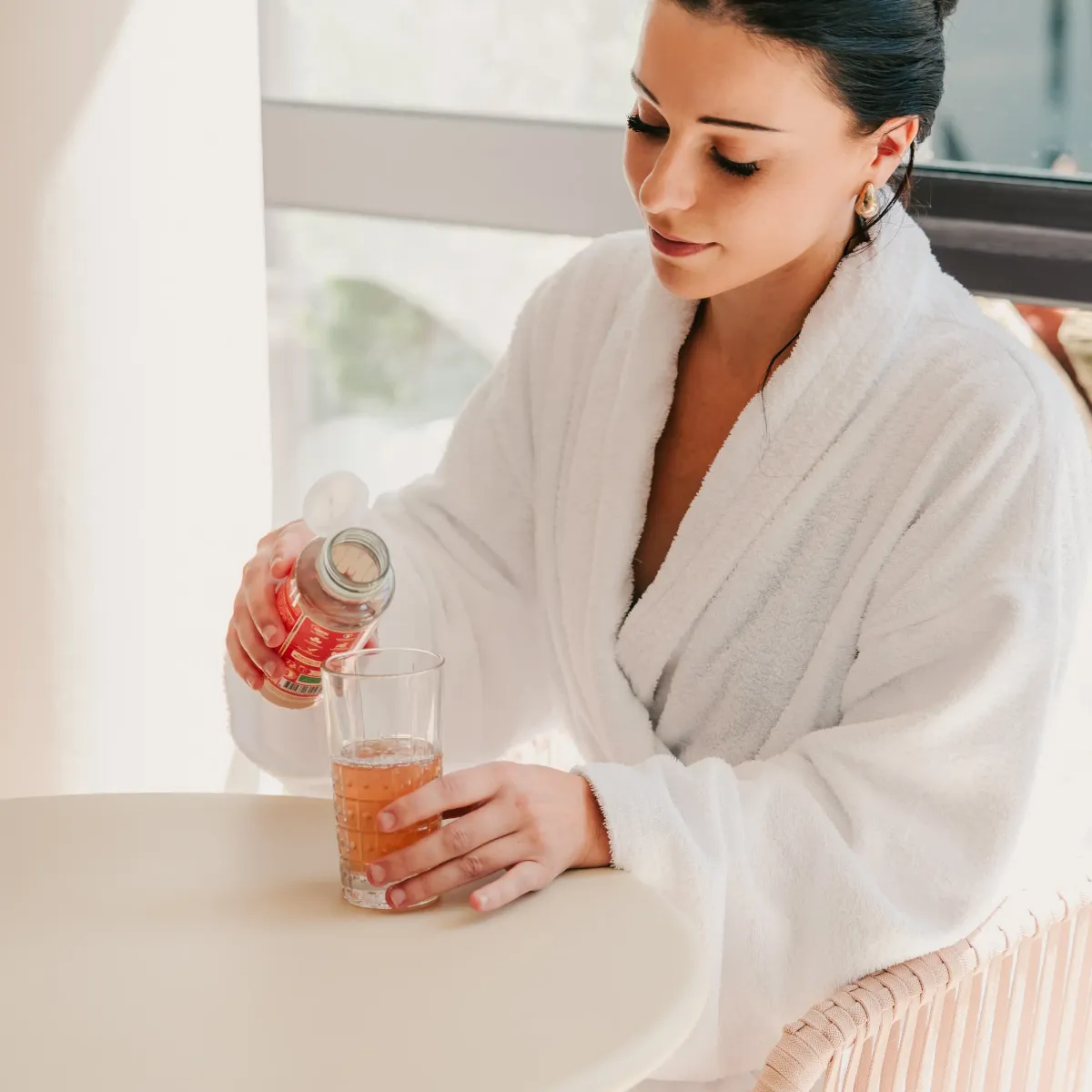 Une femme en robe de bain blanche verse un liquide dans un verre sur une table près d'une fenêtre