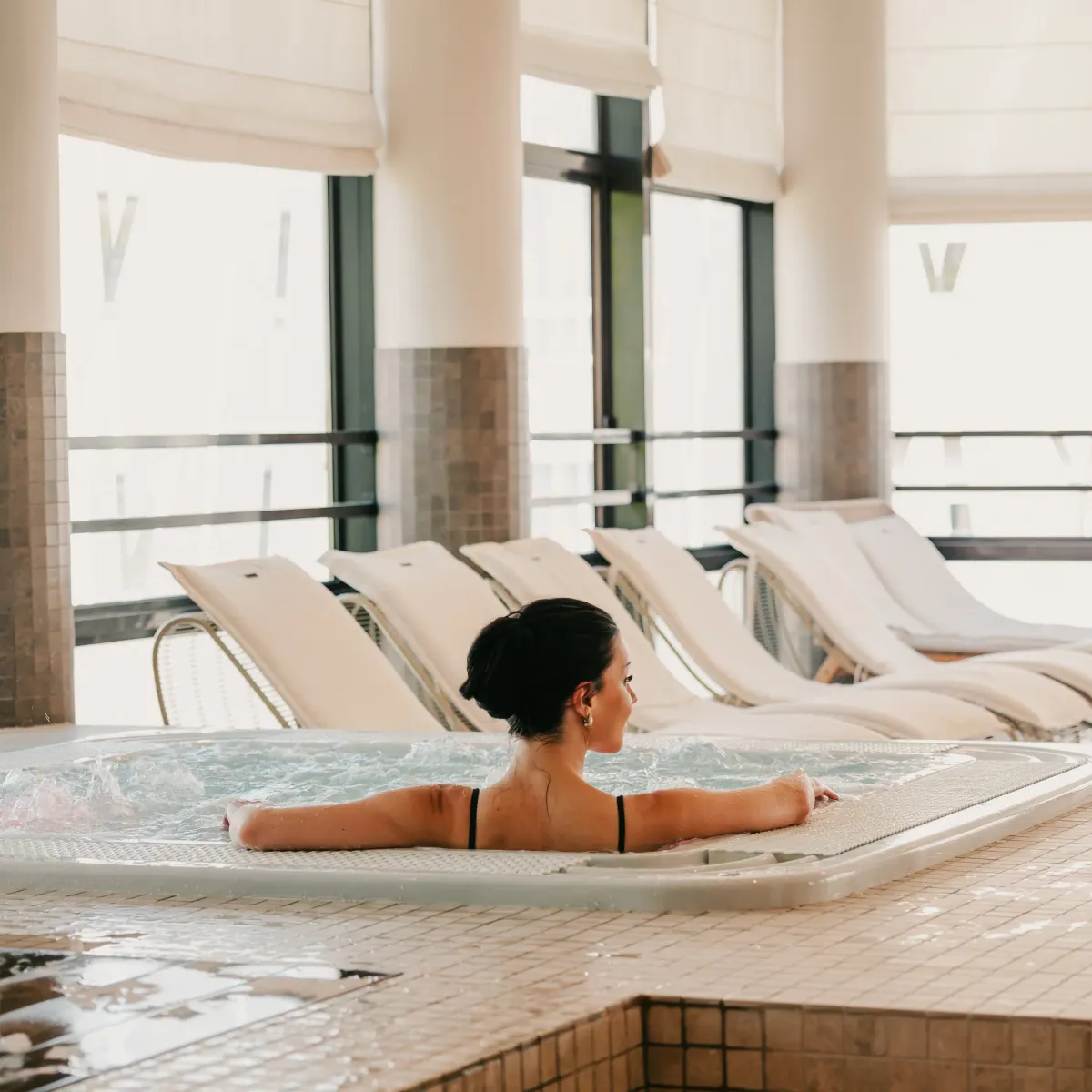 Une femme en maillot de bain noir est allongée dans un bain à remous, entourée de transats près de grandes fenêtres