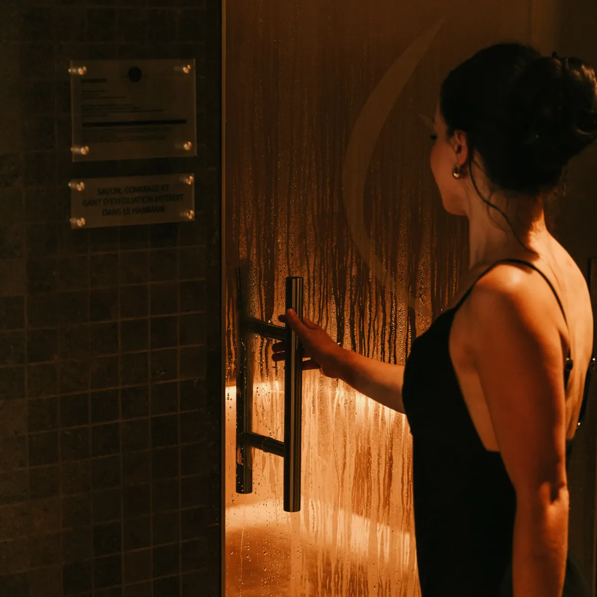 Une personne en maillot de bain noir se tient près d'une porte vitrée d'une douche, avec de l'eau coulant à l'intérieur