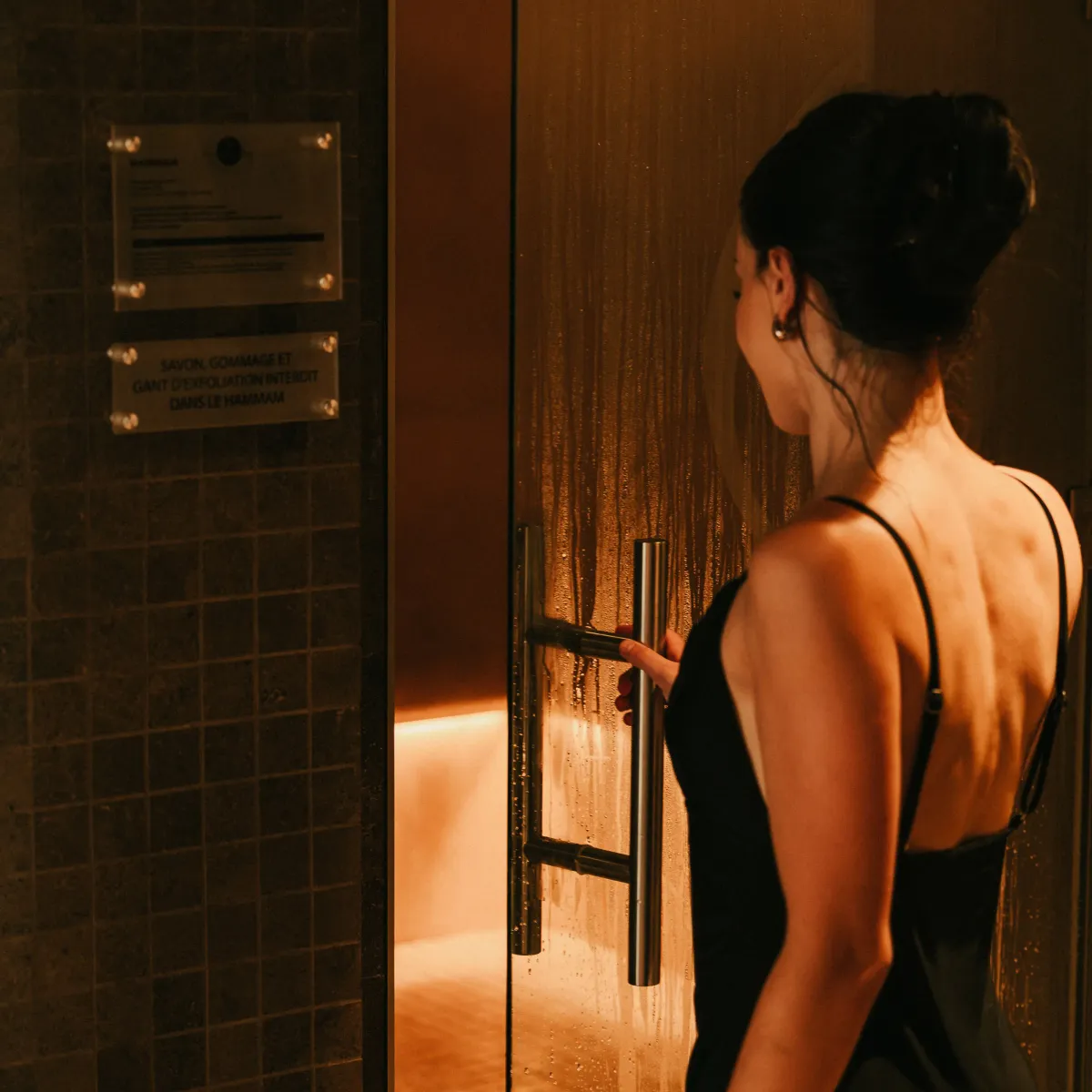 A woman in a black swimsuit opens the door of a shower cabin in a hammam.