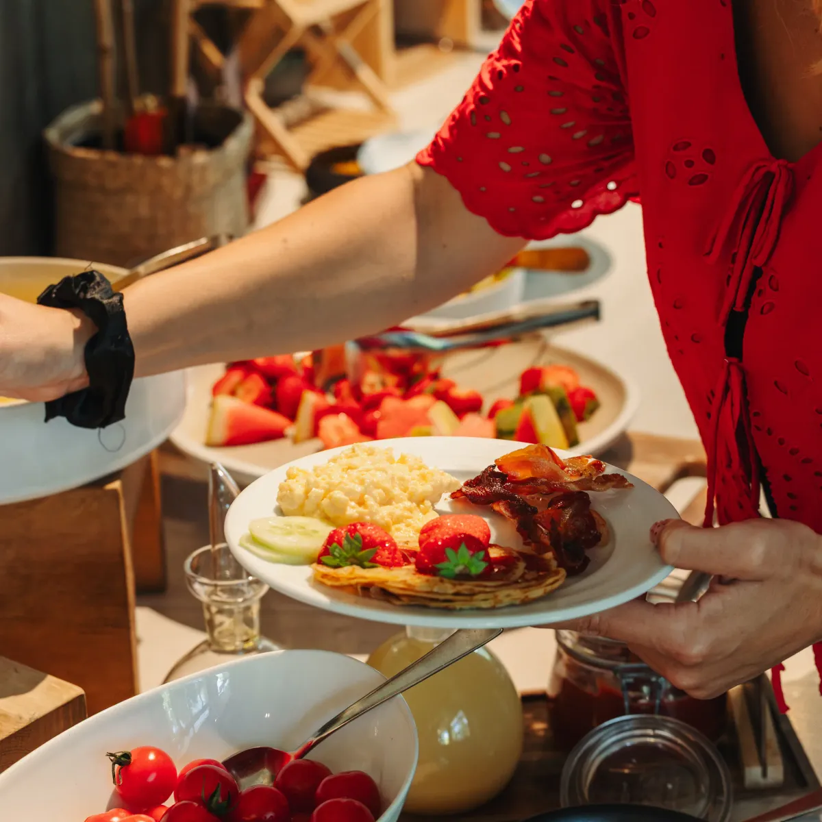 A person in a red dress is serving food onto a plate containing pancakes, fruit, and bacon, in a buffet setting.