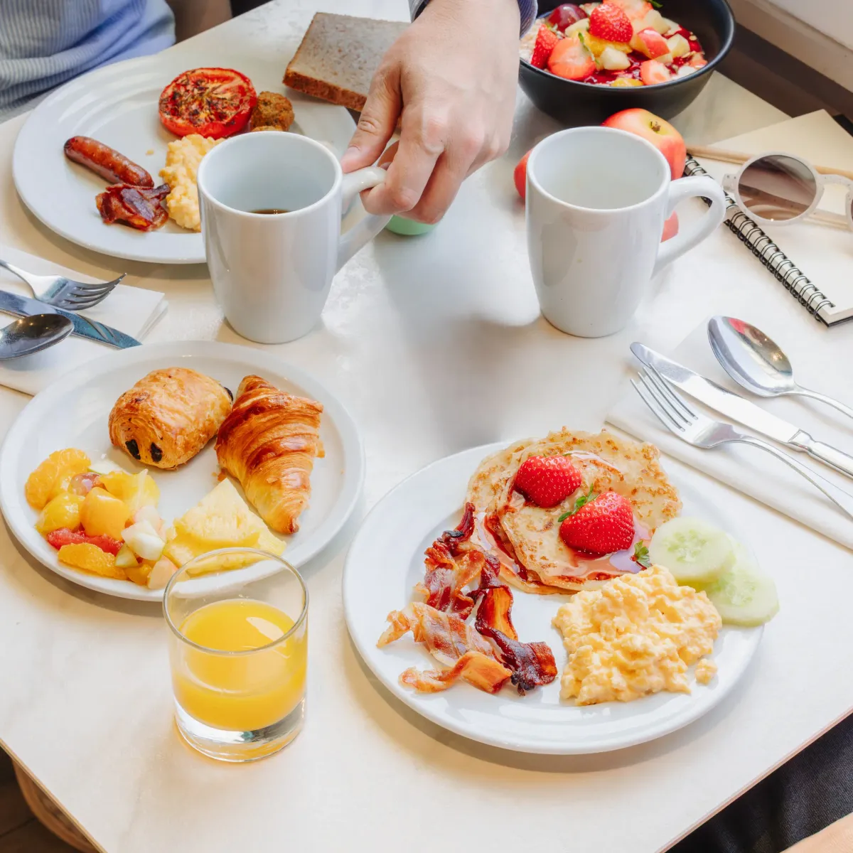 Une table avec des assiettes de petit-déjeuner, des fruits, des boissons et des couverts, une personne tenant une tasse