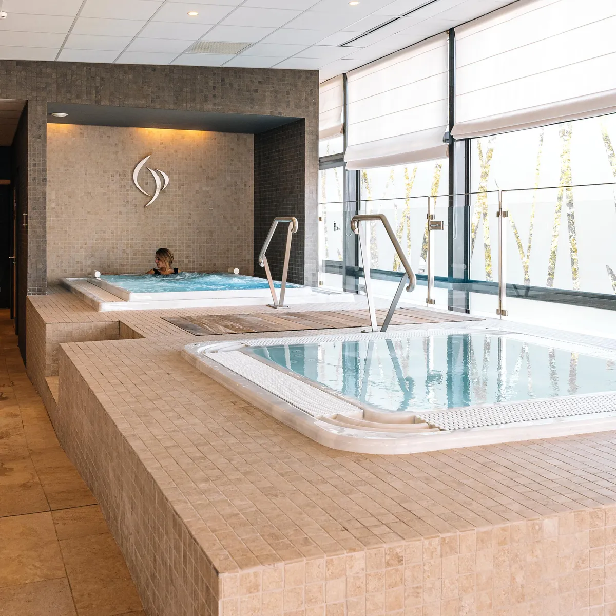 Two people are sitting in hot tubs in a bright indoor space with tiled walls and large windows.