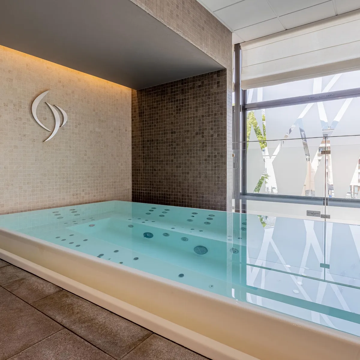 Indoor swimming pool with water jets, surrounded by tiles and large glass windows overlooking the outside.