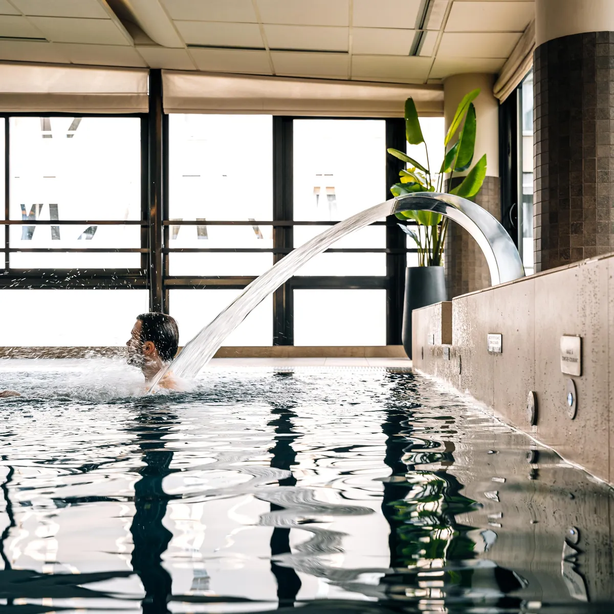 A person swims under a water jet in an indoor pool with large windows and a green plant nearby.