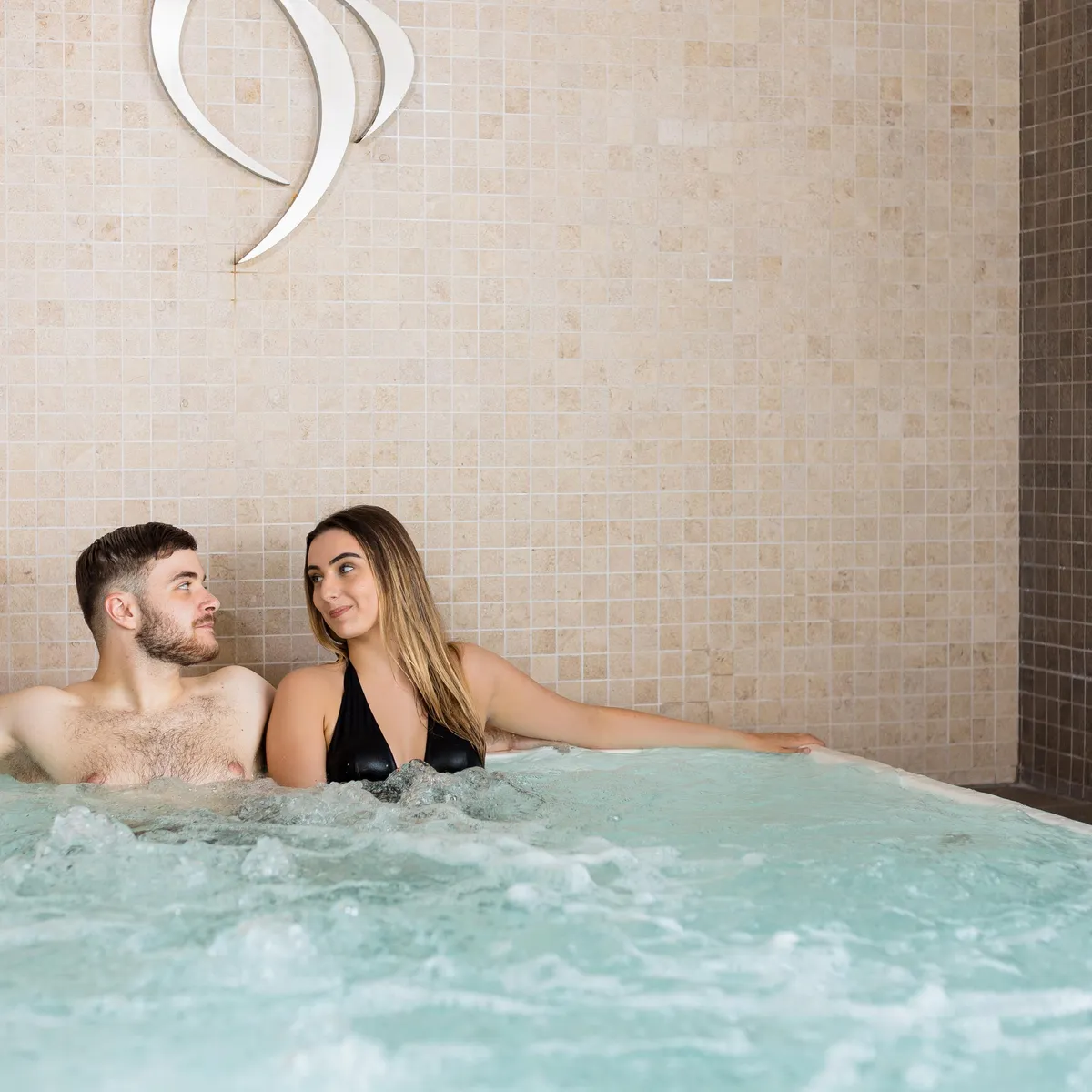 A man and a woman are sitting in a bubbling jacuzzi in front of a beige tiled wall with a white wave-shaped symbol.