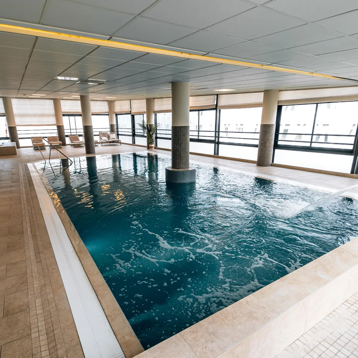 Indoor pool with columns and lounge chairs in front of large glass windows