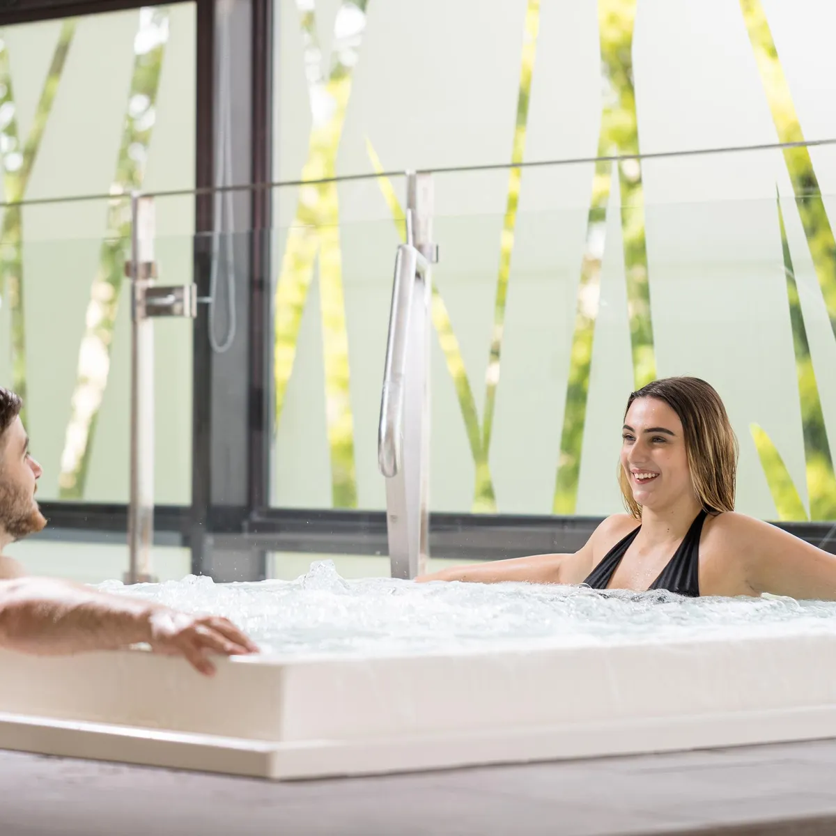 A man and a woman are sitting in a jacuzzi, surrounded by large windows with a view of green plants.