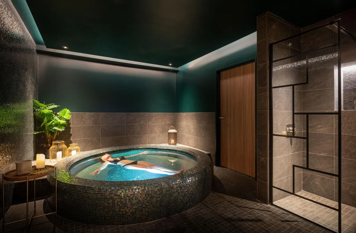 A person relaxes in a circular hot tub surrounded by mosaics, in a room with dark walls and dim lighting.